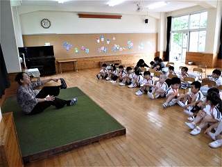 小さな台の上で先生が体育座りで体を動かす運動を園児たちに見せており、その周りの床に体育座りで座った複数の園児たちが先生の指導を聞いている様子の写真