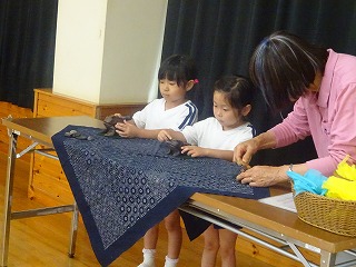 テーブルの上に広げられた紺色の模様の布の上で、体操服姿の2人の園児が、先生と一緒に工作をしている様子の写真