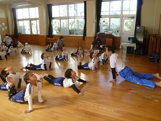 室内で体操服を着た複数の園児たちが、男性の先生の指導に合わせて、うつ伏せになり上半身を反らし足を曲げるポーズをし、柔軟体操に取り組んでいる様子の写真