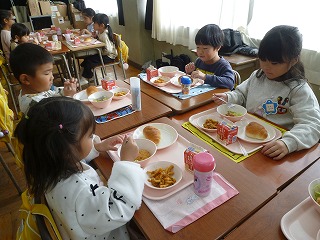 複数の園児たちが机向かい合わせでくっつけ、お盆にのったパンやおかず、牛乳などの給食を食べている様子の写真