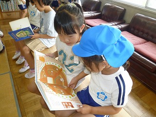 小学生の女の子が青い帽子をかぶった小さい園児に横並びに座り絵本を読んであげている様子の写真