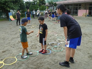 園庭の地面に等間隔に置かれた黄色や青などのフラフープの輪の中を両端からそれぞれに二人の園児が順番に飛び越えて進み、出会ったところでじゃんけんをしているのを中学の女の子が見守っている写真