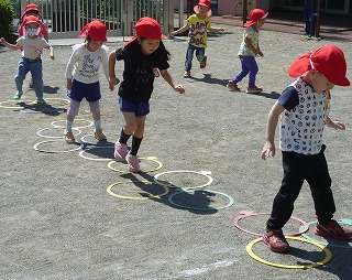 園庭で、赤い帽子をかぶった複数の園児たちが、地面に置かれた様々な色のフラフープの輪の中を、順番に踏んで進む運動遊びをしている様子の写真