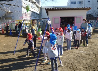 先生に見守られながら、ボールを吊り下げた青い支柱の遊具で、赤い帽子や青い帽子をかぶった複数の園児たちが一列に並び順番待ちしている様子の写真