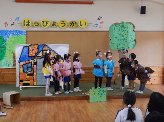 「はっぴょうかい」の看板が飾られた舞台で、園児たちが可愛らしい動物の衣装を身につけ、手作りの背景や大道具の前で劇を発表している様子の写真