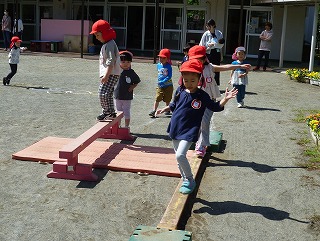 園庭で、赤い帽子をかぶった複数の園児たちが、積み上げられた赤茶色の足場と地面に置かれた平均台のような木の板の上を、バランスを取りながら慎重に渡って遊んでいる様子の写真