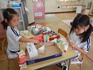 体操服を着た2人の女の子たちが、小さなテーブルの上に置かれた箱や紙コップ、様々な色の紙などの材料を使い、制作したり、組み立てたりする工作活動に取り組んでいる様子の写真