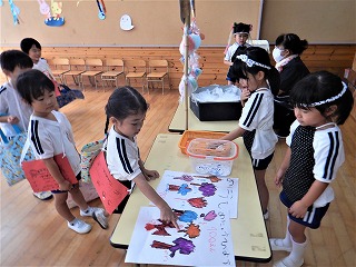 室内で体操服を着た複数の園児たちが、テーブルに広げられたイラストを見ており、机を挟んで立っている園児が鉢巻をしてお店屋さんごっこをしている様子の写真