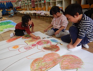 床に広げられた大きな白い紙に、3人の園児がクレヨンを使い共同で大きな絵画制作に取り組んでいる様子の写真