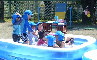 青い帽子をかぶった複数の園児たちが、屋外の園庭に設置された青いビニールプールの中に入り、水をかけ合ったりして水遊びをしている様子の写真