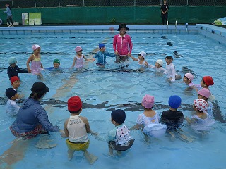 屋外の大きなプールで、水着と水泳帽を着用した複数の園児たちが、2人の先生に見守られながら、手をつないで大きな円を作り、水遊びをしている様子の写真