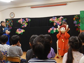 黒い背景幕と色鮮やかな造花を飾った舞台装置の前に、先生たちが大きな人形を使った人形劇を園児たちの前で上演している様子の写真