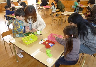 広い教室で、小さなテーブルを囲んで座る親子が、フェルトや色紙を使ってクリスマスの靴下の飾りを一緒に作っている光景を捉えた写真