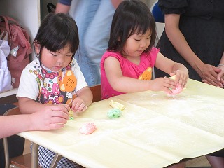 教室内で、白いシャツを着た髪の長い女の子とピンクのシャツを着た髪の長い女の子の2人が、黄色いテーブルクロスの上で粘土遊びに熱中している写真
