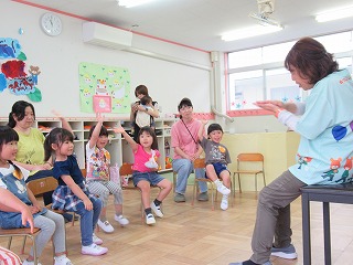 明るい保育室で、エプロン姿の女性教師が座って身振り手振りを交えながら、床に座って手を挙げたり笑顔を見せたりしている複数の園児たちと、その様子を見守る数名の保護者と思われる人々を相手に、楽しそうに活動している写真