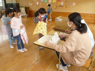 木製の床と壁の室内で、マスクを着用した先生が座る机の前に園児たちが列を作って並び、一番前の男の子が持っている袋の中を覗き込んでいる、こどもバザーの様子の写真