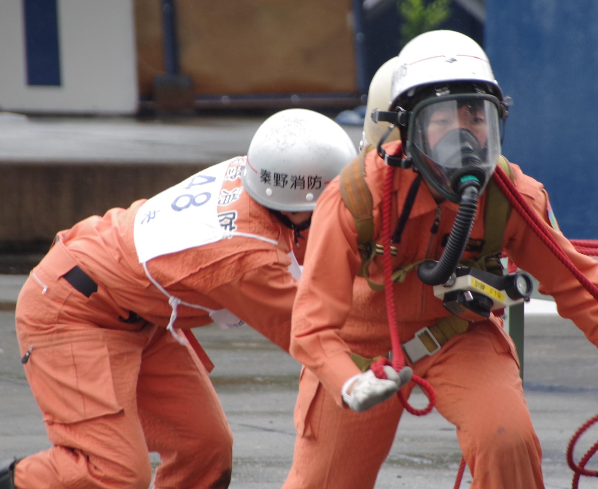 二人の救助隊員が、一人はガスマスクを装着してロープを持ち、もう一人は後ろから支えながら、真剣な面持ちで訓練に臨んでいる写真