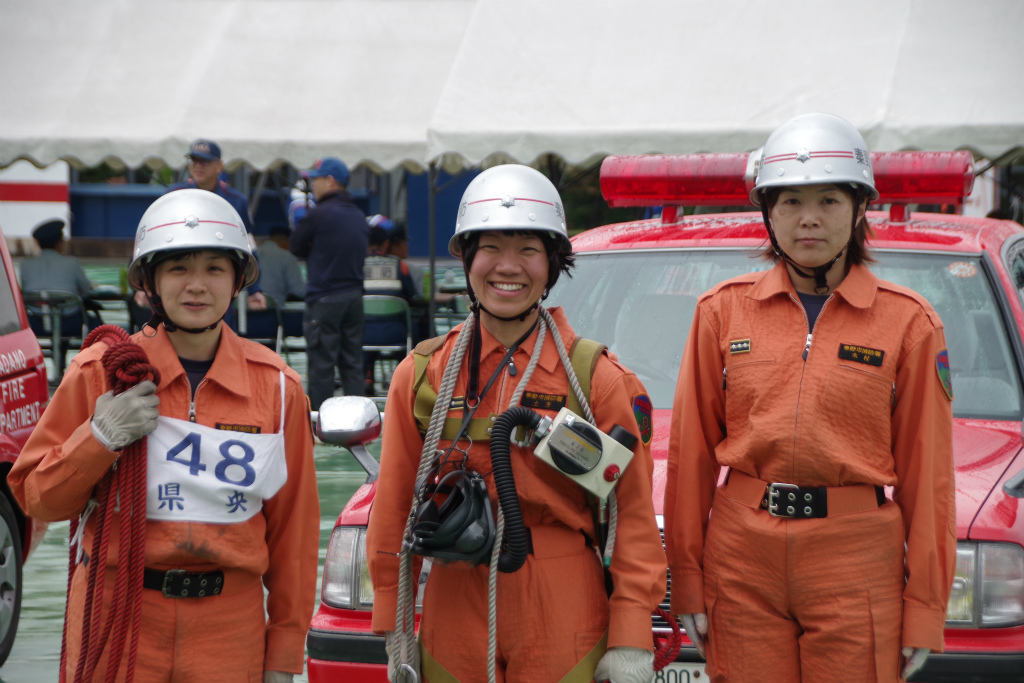 赤い車を背景に、消防救助の訓練に参加した3人の女性隊員が、真ん中の1人は笑顔で、他2人は真剣な表情で並んで写っている記念写真