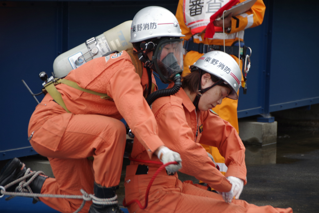 背中にボンベを背負い、ガスマスクを装着した救助隊員が、要救助者役の隊員の腰にロープを結んでいる様子を写した写真