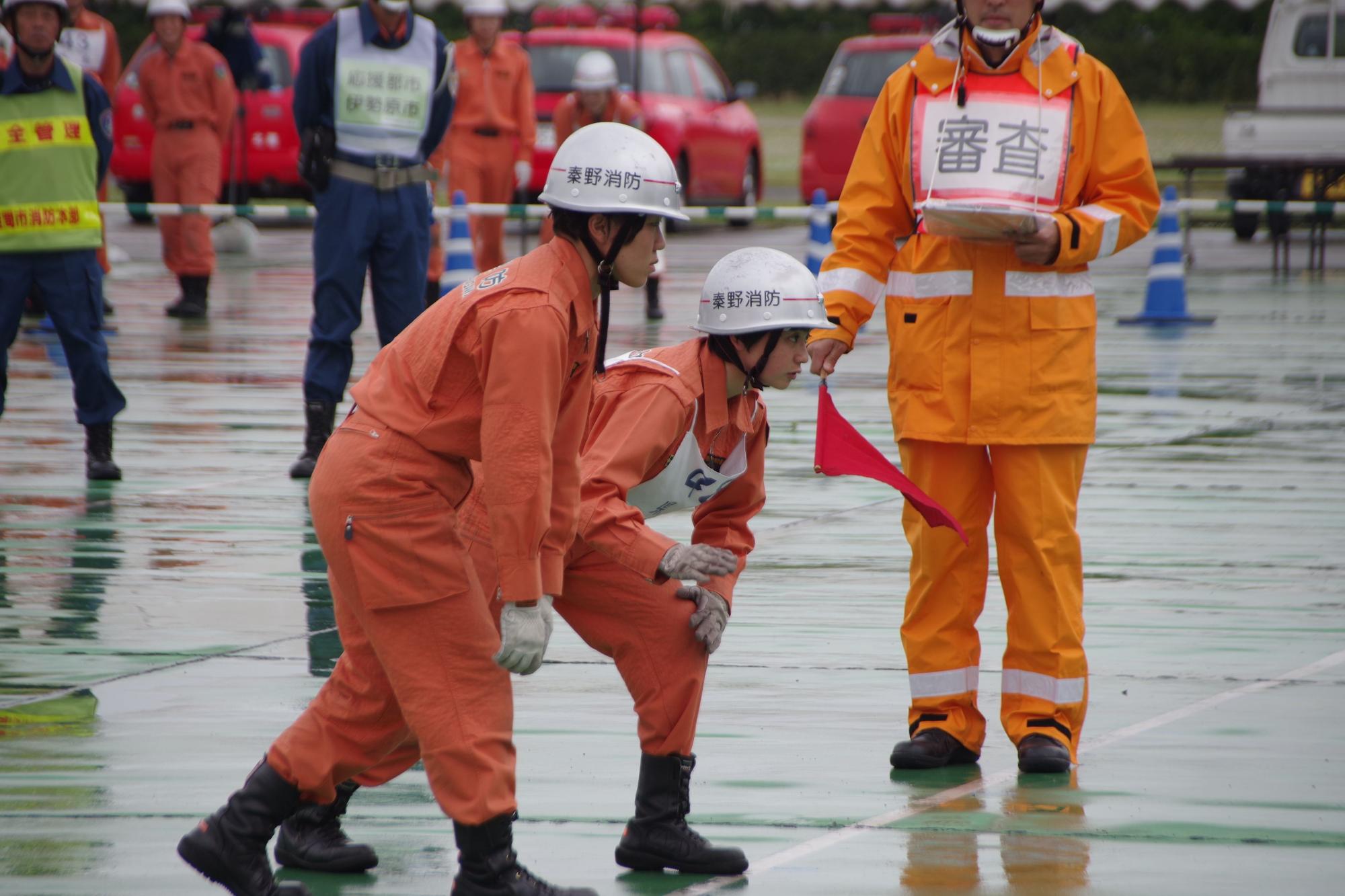 審判が赤い旗を持っている前で、2人の女性救助隊員が訓練の合図を待って、スタートラインに身構えている写真
