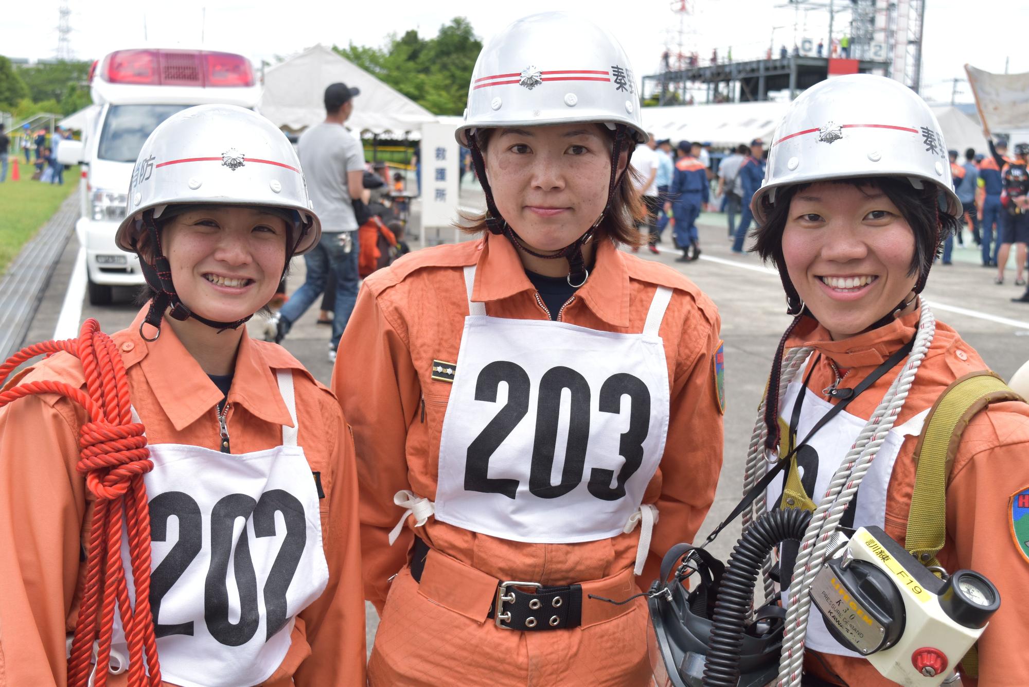 屋外で、救急車や白いテントを背景に、オレンジ色のレスキュー服にゼッケンをつけ、銀色のヘルメットを着用した3名の女性隊員が、カメラに向かって笑顔を見せている写真