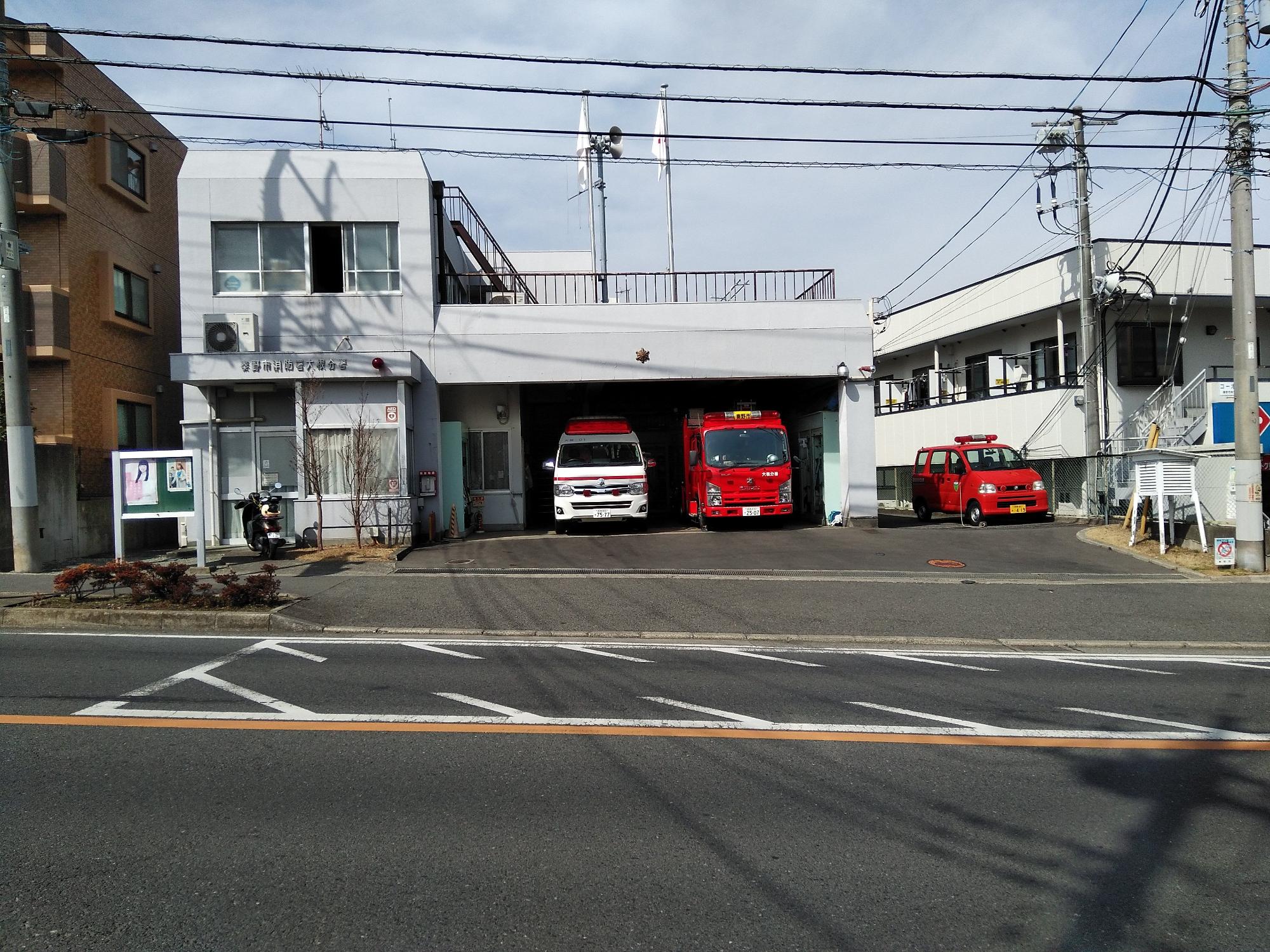 正面中央に2台の緊急車両が待機する車庫と、右側の屋外に1台の軽消防自動車が停車し、屋上に手すりのついたバルコニーを備えた2階建ての消防署大根分署の外観写真