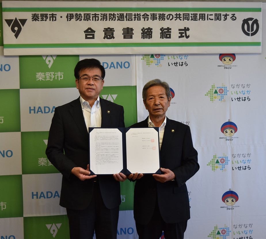 「秦野市・伊勢原市消防通信指令事務の共同運用に関する合意書締結式」と書かれた横断幕の前で、スーツ姿の市長と男性が合意書を中央に持ち、まっすぐカメラを見て立っている記念写真