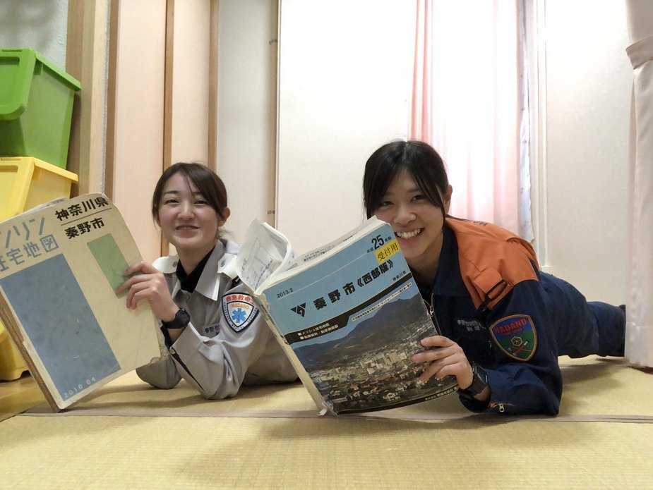 救急服の女性とレスキュー服の女性が、それぞれ「住宅地図 秦野市」と「秦野市（西部編）」の厚い本を広げてカメラに向かって笑顔を見せている、現場活動前の勉強の様子の写真