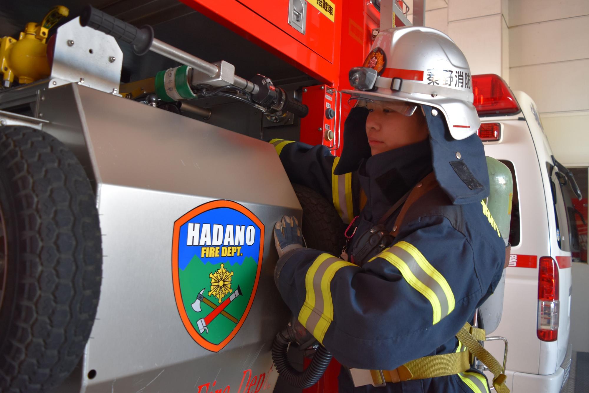 消火活動用の装備を完全に着用し、銀色のヘルメットをかぶった女性消防隊員が、秦野市消防本部のエンブレムが貼られた消防車の側面で作業している様子の写真