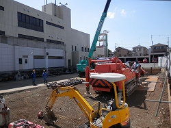 クレーン車やショベルカーなどの複数の重機と、ヘルメットを被った複数の作業員が立っている基礎工事中の建設現場の写真