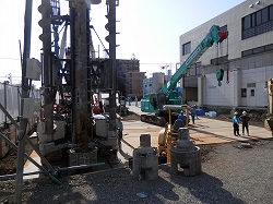 掘削用の大きなドリルやクレーン車などの重機が配置されており、ヘルメットを被った複数の作業員が作業している建設現場の写真