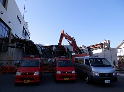 消防署の一部がショベルカーで取り壊され、その前には複数の小型消防車やバンが駐車している解体工事現場の写真