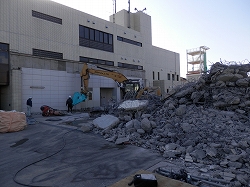 解体中の消防署で、ショベルカーが瓦礫を運んでおり、その手前には複数の作業員や、がれきの山が写っている解体工事現場の写真