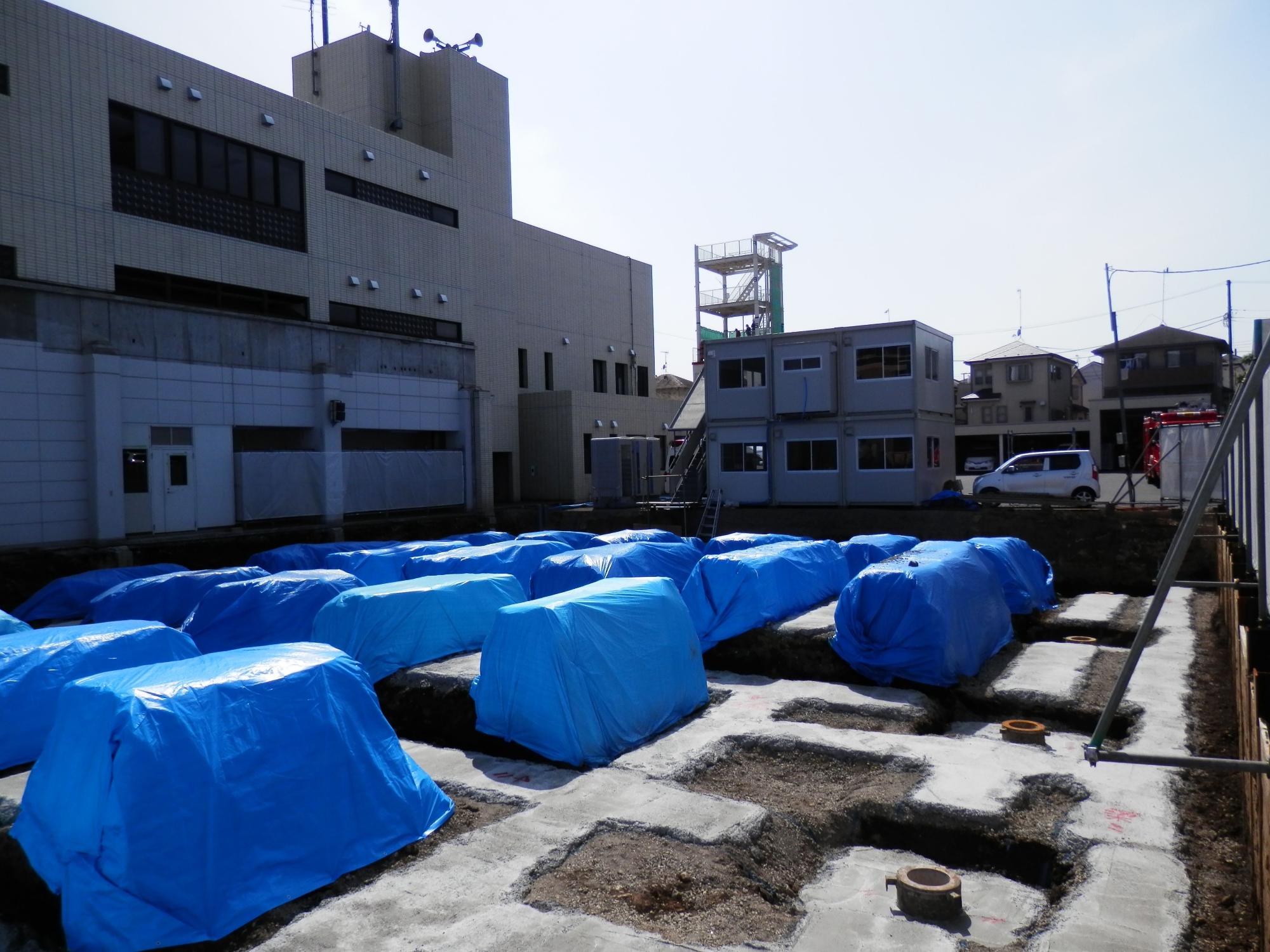 工事中の建物の基礎部分で、複数の土台が作られており、それらが青いビニールシートで覆われている建設現場の写真