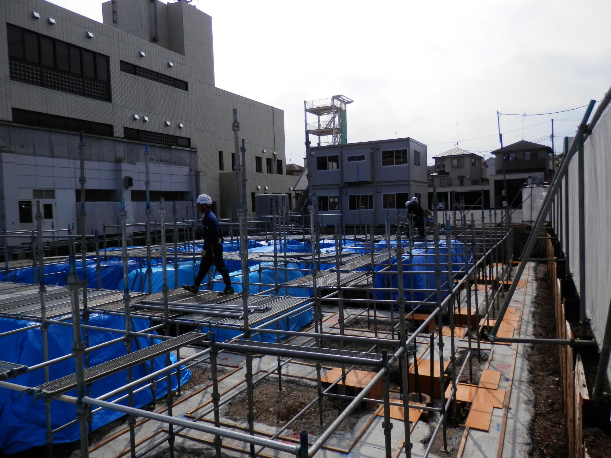 グリッド状に多数の鉄筋が配置されコンクリート打設前の準備が進む新築の基礎工事現場を、手前の足場板越しに捉えた写真