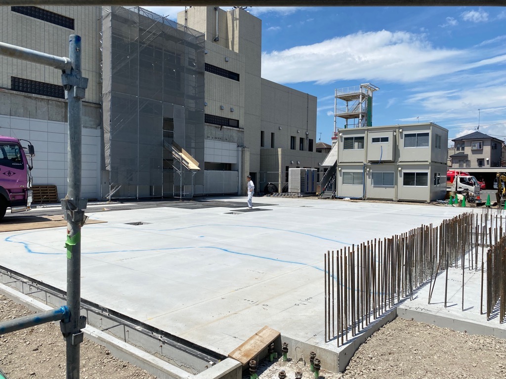 奥にプレハブの仮設事務所が設置されている建設中の建物の手前に、鉄筋が多数突き出たコンクリートの基礎部分が広がっている写真
