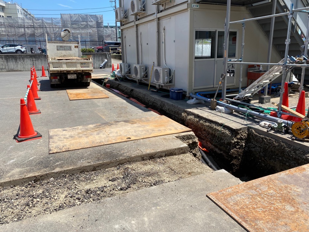 アスファルトの地面が掘り起こされ、配管やケーブルがむき出しになっており、その横に工事用のコーンや仮設の建物が設置されている建設現場の写真
