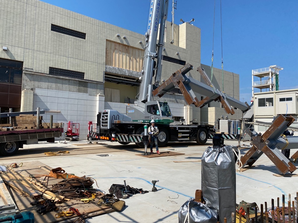 複数の建設機械と資材が置かれた工事現場で、大きなクレーンが鉄骨の資材を吊り上げている様子を写した写真