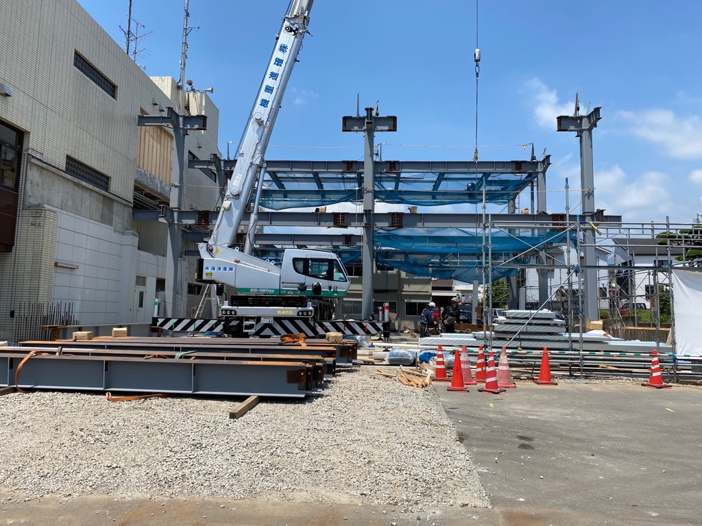 クレーン車が鉄骨を吊り上げており、地面には資材が山積みになっている建設中の工事現場の写真