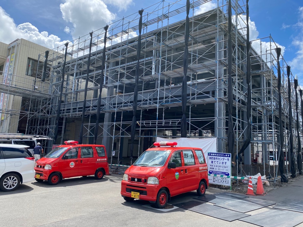 白いシートがかけられ、足場が組まれた建物の前で、赤くて小さな消防車が2台駐車している建設中の消防署の写真