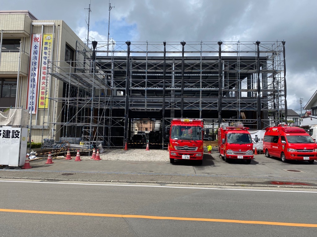 骨組みがむき出しで、足場が組まれた建物の前に、複数の赤い消防車が駐車している、建設中の消防署の写真