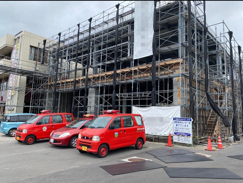建物の周りを工事の足場と黒いシートが覆っており、その手前には赤い消防車が3台駐車されている、建設中の消防署の写真