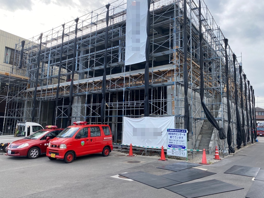 工事用の看板が置かれて、前には2台の消防車が駐車しており、工事の足場と黒いシートで覆われた建物の写真