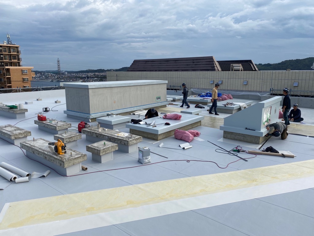 たくさんの機器が置かれ、複数の作業員が屋上の防水工事をしている写真