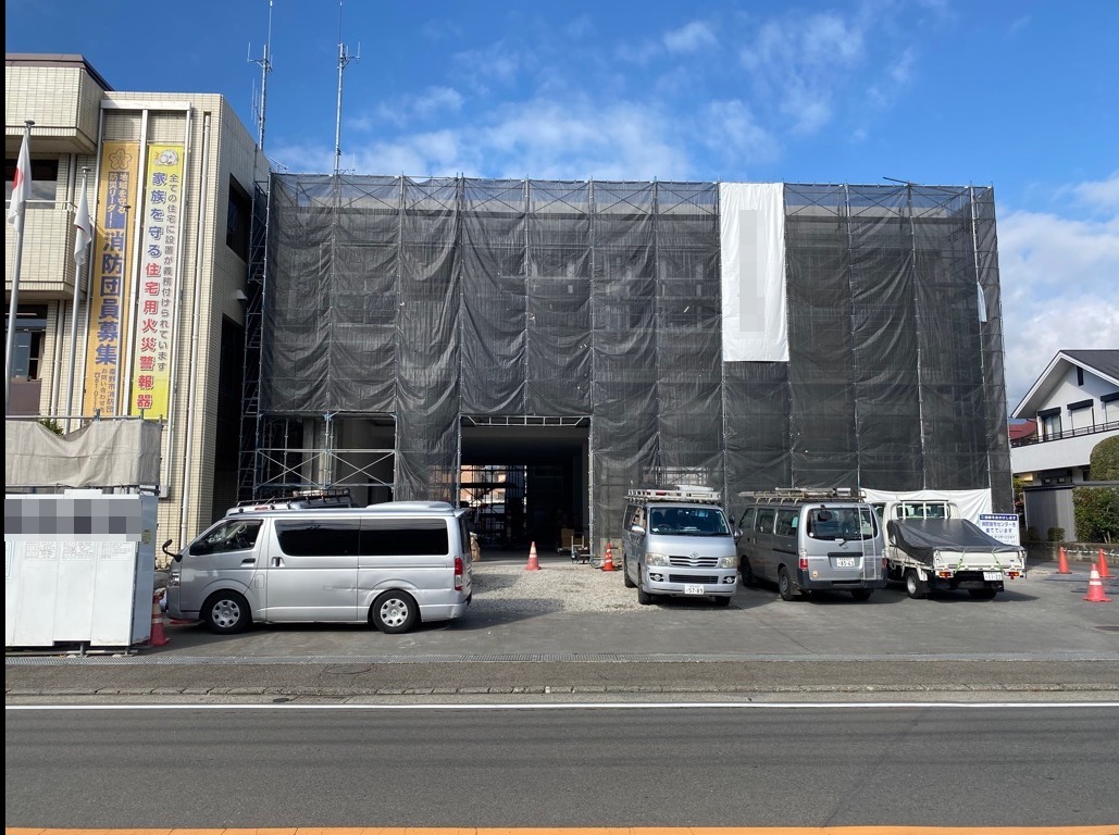白い建物全体に足場と黒い養生ネットがかけられており、複数の車が駐車している、改修工事中の消防署の写真