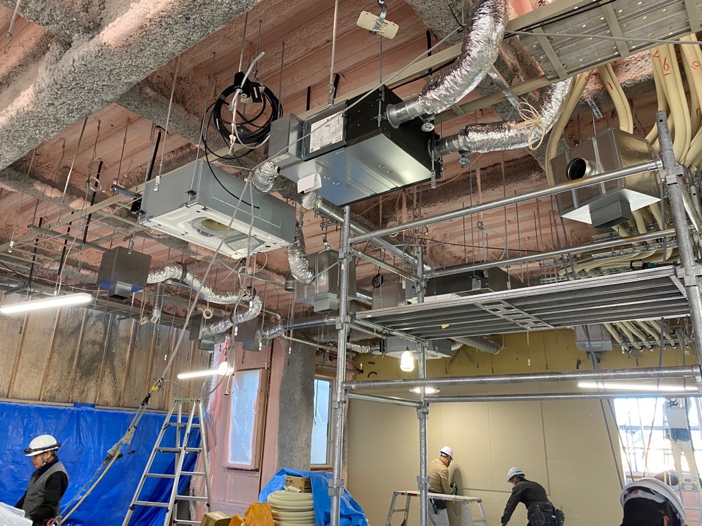 たくさんの空調ダクトや配管がむき出しになった、建設中の建物の天井で、ヘルメットを被った複数の作業員が作業をしている写真