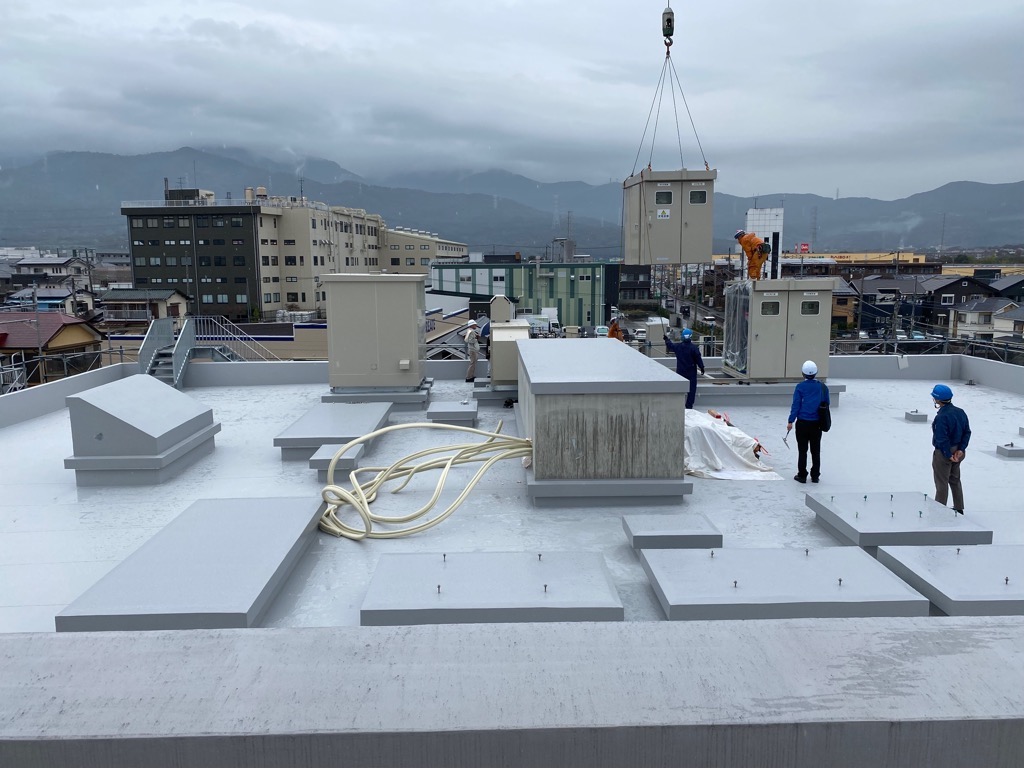 ビルの屋上に複数の作業員がおり、クレーンを使って大きな機器を吊り上げている建設現場の写真