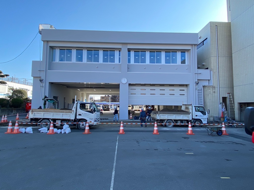 建物の前で複数の作業員が土砂を運んでおり、工事用のコーンやトラックが並んでいる建設中の消防署の写真
