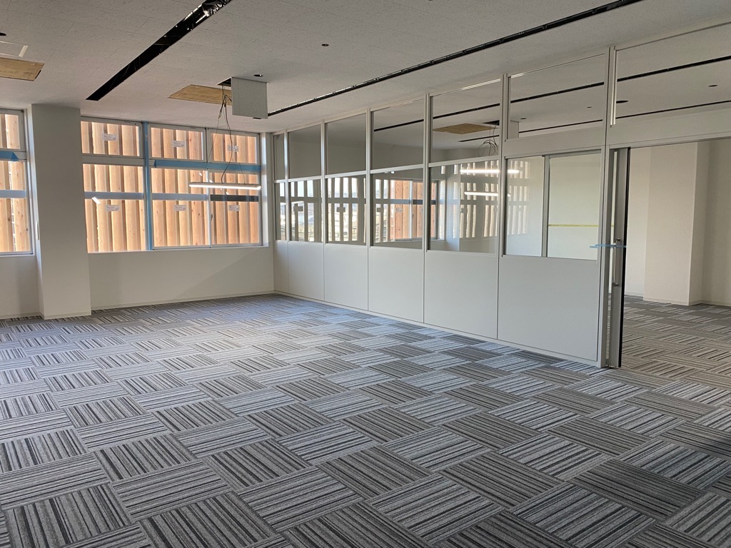 白い間仕切りとガラス扉が設置され、床には白と黒の幾何学模様のカーペットが敷かれた事務室の写真