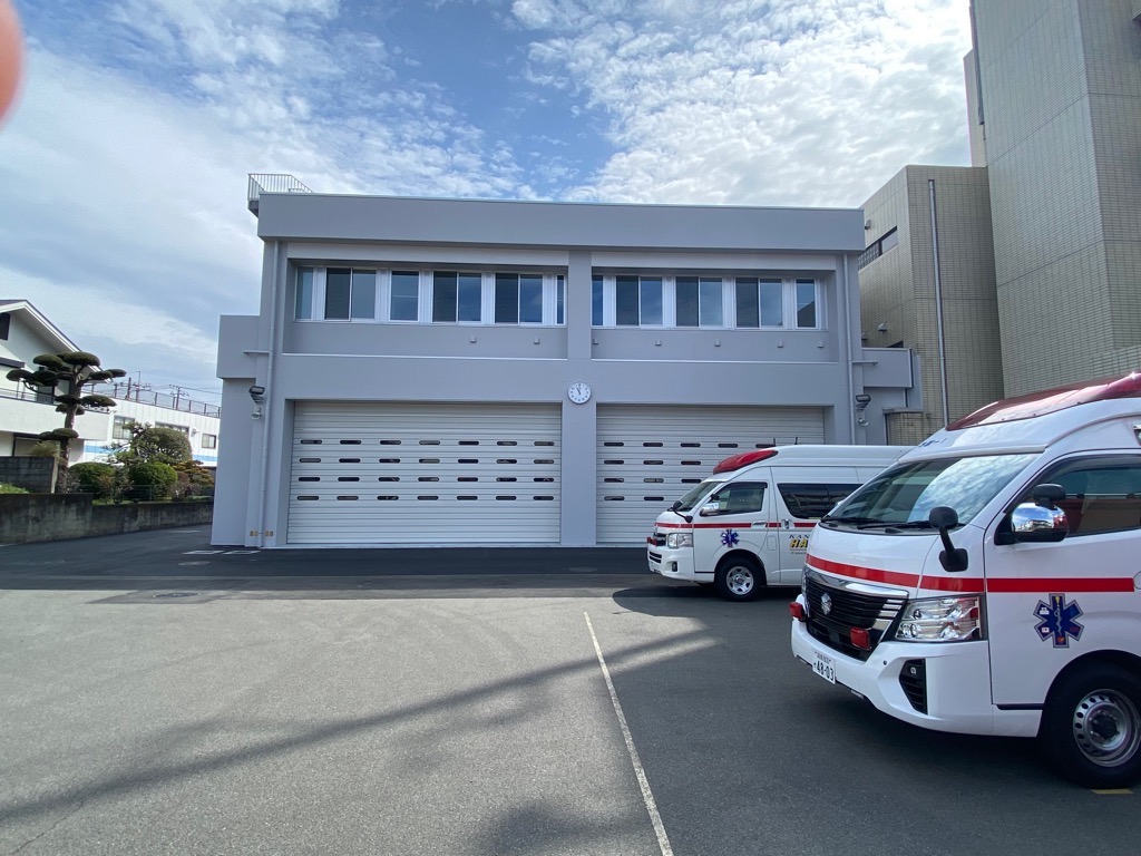グレー色の建物とシャッター付きの車庫の前に、2台の白い救急車が駐車している消防署の写真